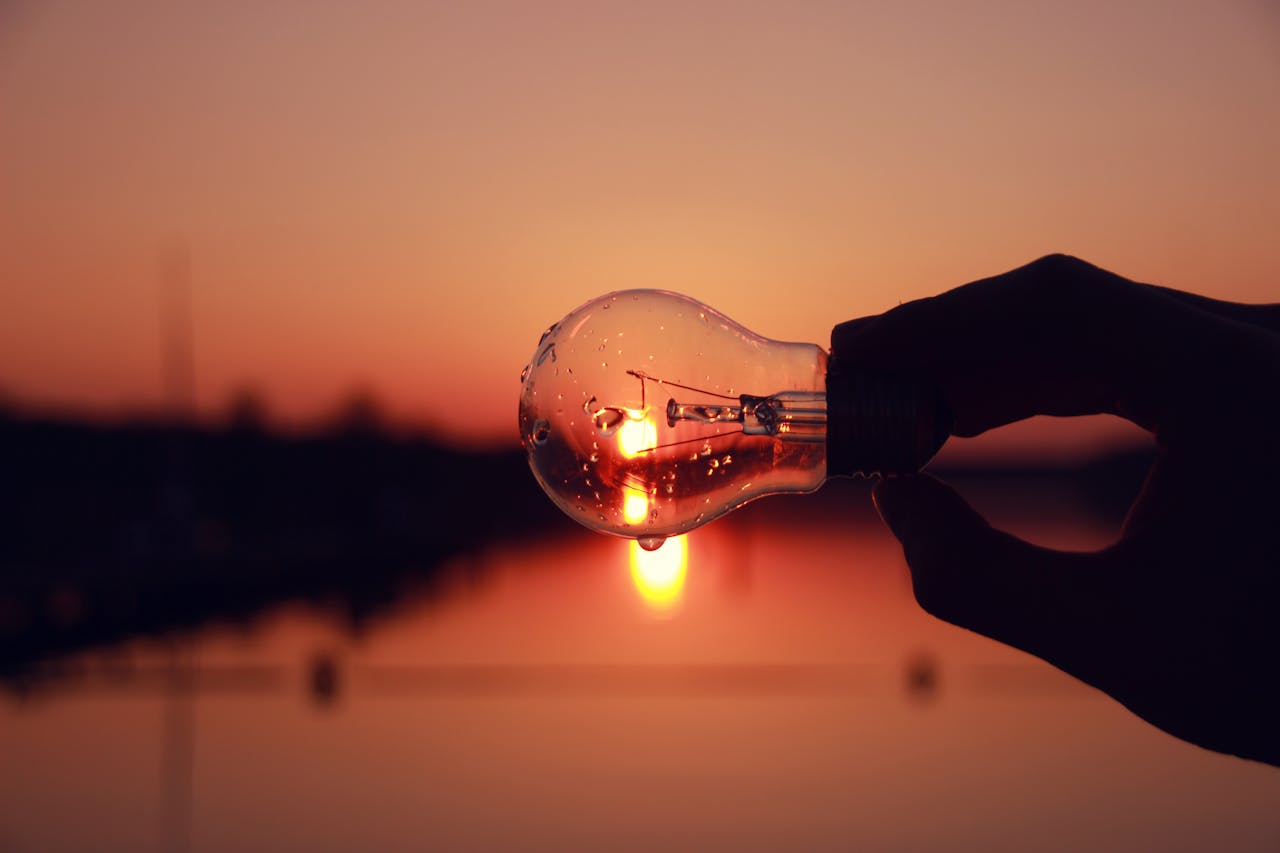 Silhouette of a hand holding a light bulb against a stunning sunset over a lake, blending creativity and nature.