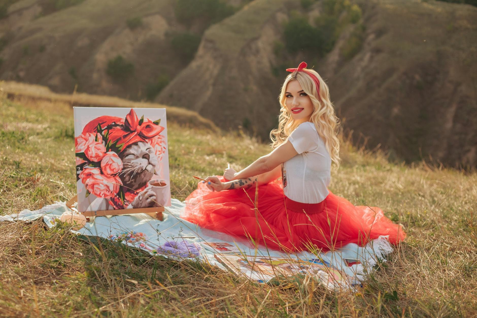 A smiling woman in a red tutu skirt paints in a sunny London meadow.