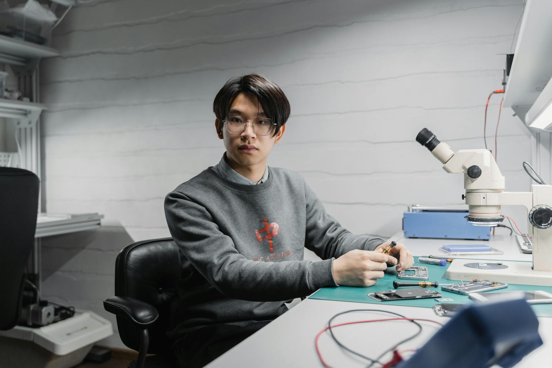Asian man working with soldering tools and microscope on electronics in a lab setting.
