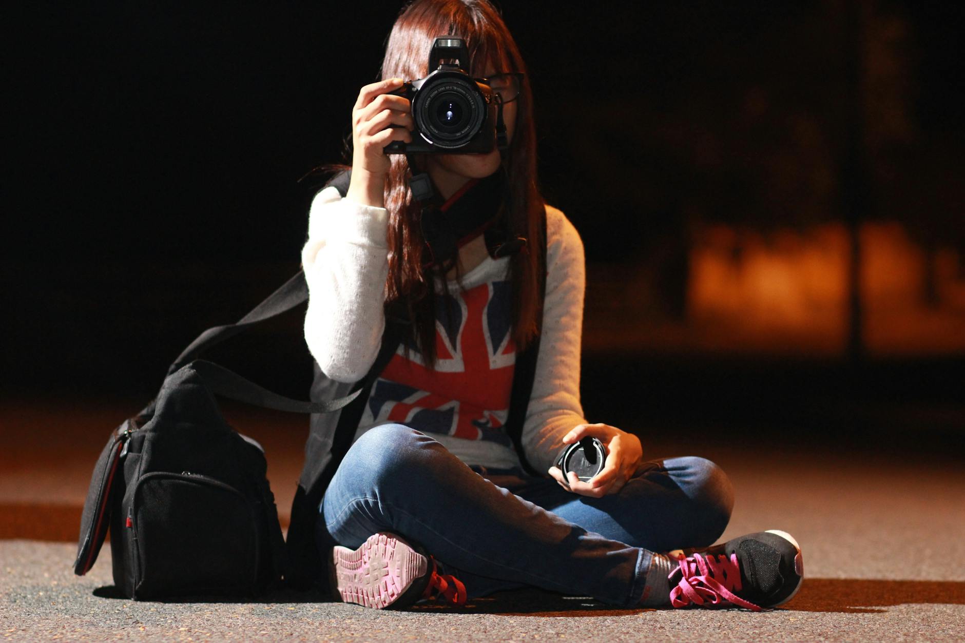 A woman sitting outdoors at night, capturing a photo with a DSLR camera, showcasing urban exploration.