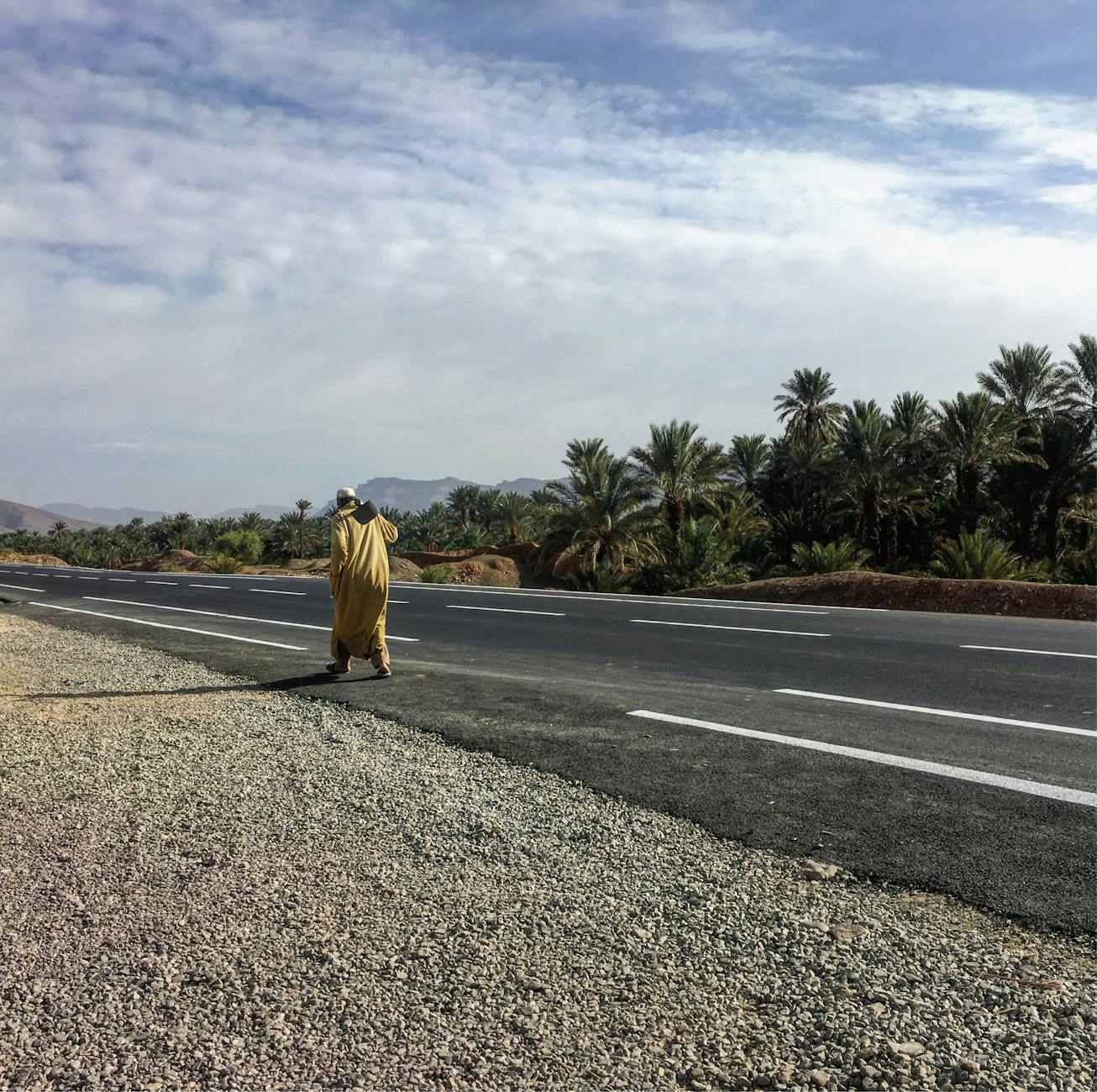 A man in traditional attire walking on a road in Ouled Otmane, Morocco, with palm trees in the background.